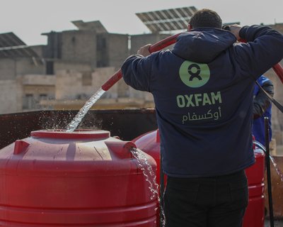 A person wearing an Oxfam jacket is shown with a hose with running water coming out of it into a tank outside in Aleppo.